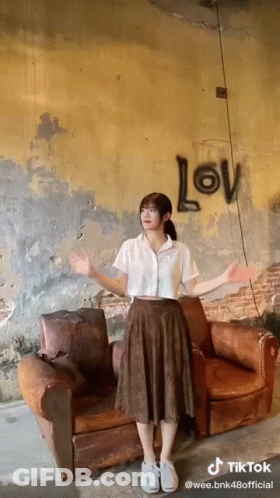 A Girl Is Standing In Front Of A Couch With The Word Love Written On The Wall Behind Her GIF