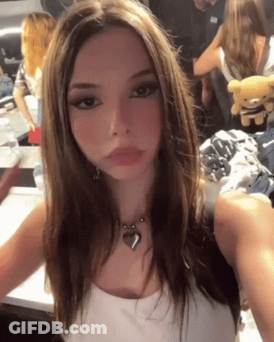 A Girl Is Taking A Selfie With A Teddy Bear In The Background While Wearing A Heart Necklace . GIF