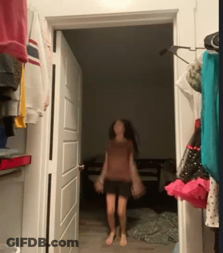 A Girl Is Walking Through A Doorway In A Room With Clothes Hanging On The Wall . GIF