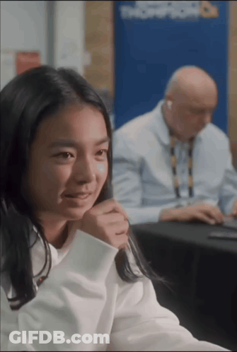 A Girl Sits At A Table With A Man Behind Her And A Sign That Says Troop Room On It GIF