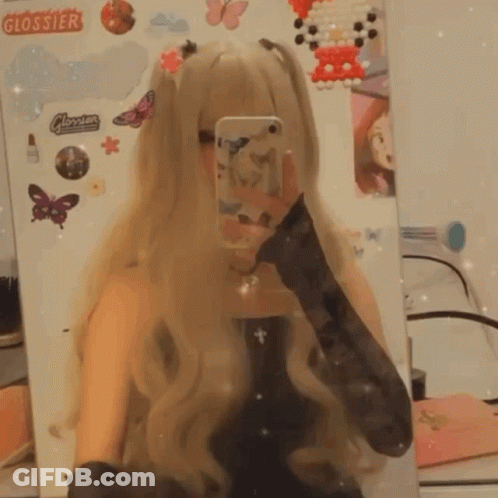A Girl Taking A Selfie In Front Of A Mirror With Stickers On The Wall That Say Glossier GIF