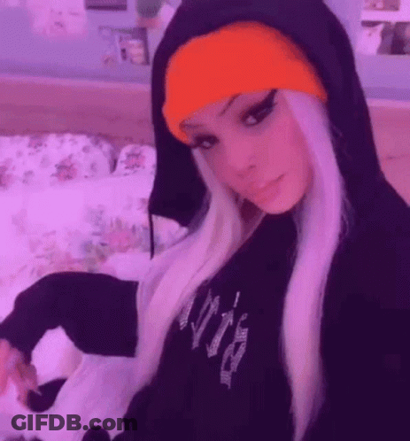 A Girl Wearing A Black Hoodie And An Orange Beanie Is Sitting On A Bed . GIF