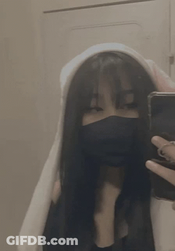 A Girl Wearing A Mask And A Hoodie Is Taking A Selfie With Her Phone . GIF