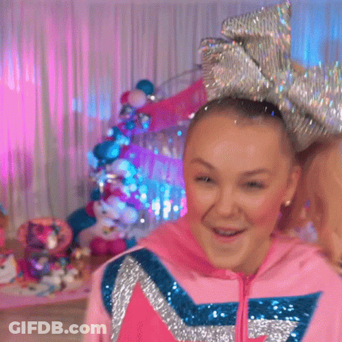 A Girl Wearing A Pink Hoodie And A Large Bow In Her Hair Is Standing In A Room . GIF