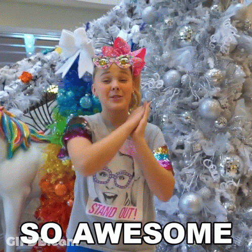 A Girl Wearing A T-Shirt That Says " Stand Out " Is Clapping In Front Of A Christmas Tree GIF