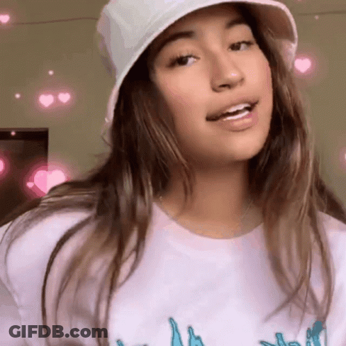 A Girl Wearing A White Hat And A White T-Shirt Is Smiling And Looking At The Camera . GIF