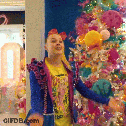 A Girl Wearing A Yellow Shirt That Says ' Jojo ' On It Is Dancing In Front Of A Christmas Tree GIF