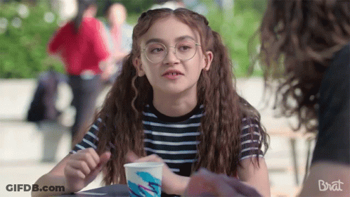 A Girl Wearing Glasses Is Sitting At A Table With A Cup Of Ice Cream In Front Of Her . GIF