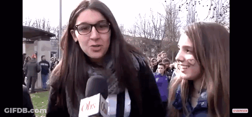 A Girl Wearing Glasses Is Talking Into A Microphone While Another Girl Has A Heart Drawn On Her Face . GIF