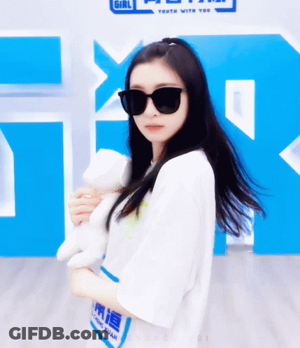 A Girl Wearing Sunglasses Is Holding A Stuffed Animal In Front Of A Sign That Says Girl Youth With You GIF