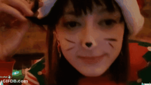 A Girl With A Cat Face Painted On Her Face And A Santa Hat GIF