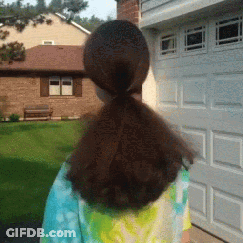 A Girl With A Ponytail Is Standing In Front Of A Garage Door . GIF