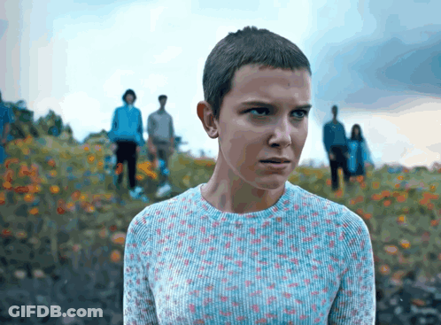 A Girl With A Very Short Haircut Is Standing In A Field Of Flowers GIF