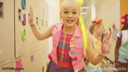 A Girl With A Yellow Bow In Her Hair Is Standing In A Hallway With Other Girls GIF