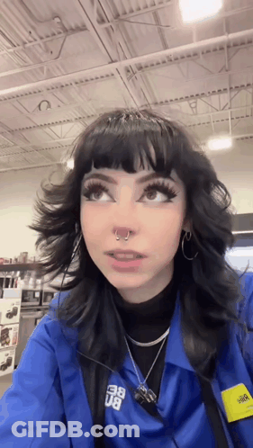 A Girl With Black Hair And A Nose Ring Is Wearing A Blue Jacket And Earrings . GIF