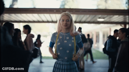 A Girl With Blonde Hair And A Blue Shirt Is Standing In A Hallway GIF