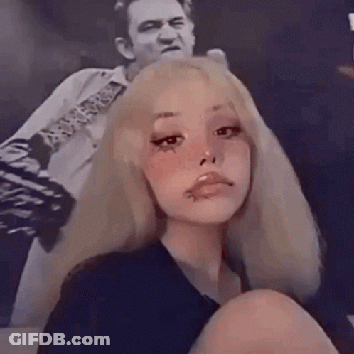 A Girl With Blonde Hair And Freckles Is Sitting In Front Of A Picture Of A Man Playing A Guitar . GIF