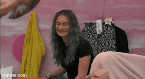 A Girl With Blue Hair Is Smiling In Front Of A Pink Wall With Clothes Hanging On It GIF