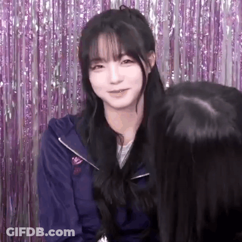 A Girl With Long Black Hair And Bangs Is Standing In Front Of A Purple Curtain . GIF