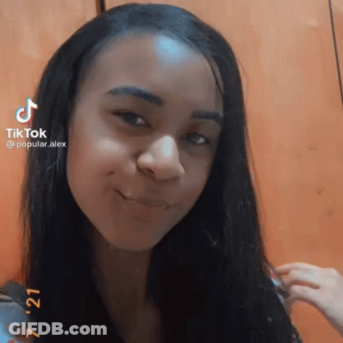 A Girl With Long Black Hair Is Smiling And Looking At The Camera While Holding Her Hair . GIF