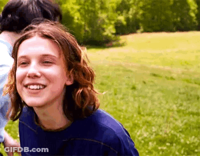A Girl With Long Hair Is Smiling While Standing In A Field . GIF