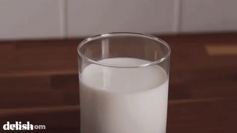 A Glass Of Milk Is Sitting On A Wooden Table With The Word Delish On The Bottom . GIF