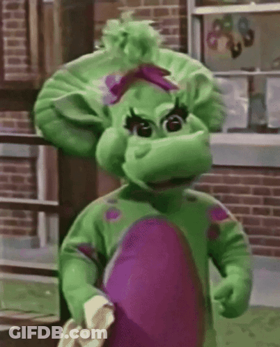 A Green And Purple Dinosaur Mascot Standing In Front Of A Brick Building . GIF