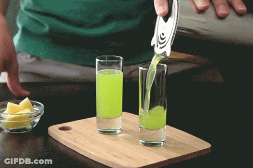 A Green Liquid Is Being Poured Into Two Shot Glasses On A Cutting Board GIF