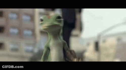 A Green Lizard Is Standing In Front Of A Building And Looking At The Camera GIF