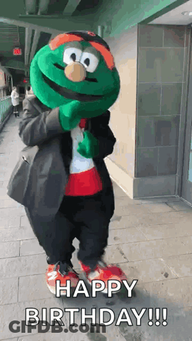 A Green Mascot Is Standing On A Sidewalk And Says Happy Birthday ! GIF