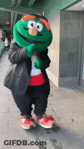 A Green Mascot Is Standing On A Sidewalk Wearing A Suit And A Hat . GIF