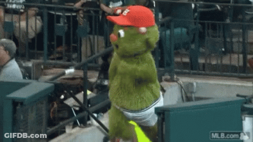 A Green Mascot Wearing A Red Hat And Shorts Is Dancing In The Stands At A Baseball Game .. GIF