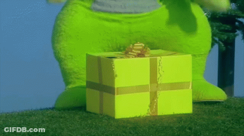 A Green Stuffed Animal Standing Next To A Yellow Gift Box With A Gold Bow GIF