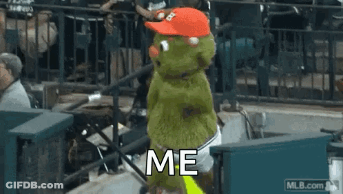 A Green Stuffed Animal Wearing A Red Hat Is Standing In A Stadium . GIF