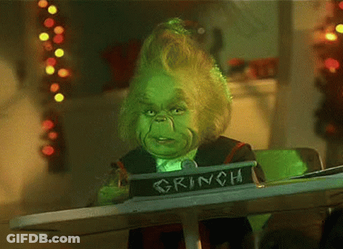 A Grinch Is Sitting At A Table With A Sign That Says Grinch On It . GIF