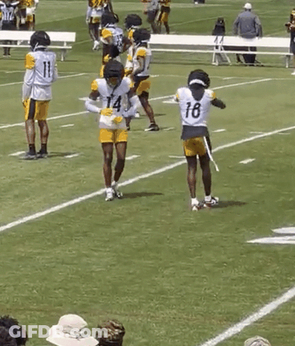 A Group Of Football Players Are Practicing On A Field While A Man In A Yellow Shirt Watches . GIF