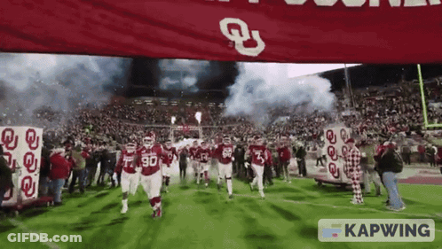 A Group Of Football Players Are Running On A Field With A Banner That Says Kapwing In The Foreground GIF