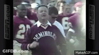 A Group Of Football Players Are Standing Around A Man Wearing A Sweater That Says ' Miami ' On It . GIF