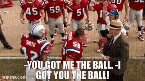 A Group Of Football Players Are Standing On A Field Talking To A Man In A Suit . GIF