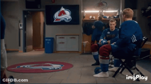 A Group Of Hockey Players Are Sitting In A Locker Room With A Tv Screen Behind Them That Says Avalanche On It GIF