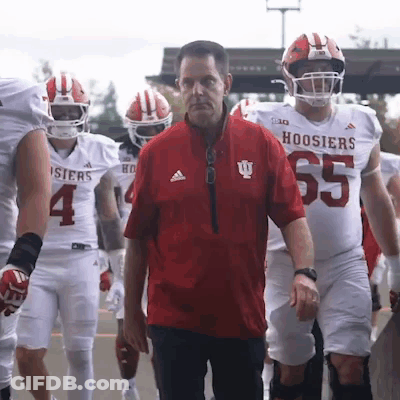A Group Of Hoosier Football Players Are Walking Towards A Man In A Red Jacket GIF