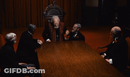 A Group Of Men Are Sitting Around A Table With A Man Standing In The Middle GIF