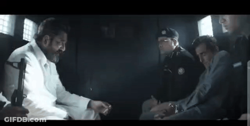 A Group Of Men Are Sitting In A Dark Room Talking To Each Other . One Of The Men Is Holding A Gun . GIF