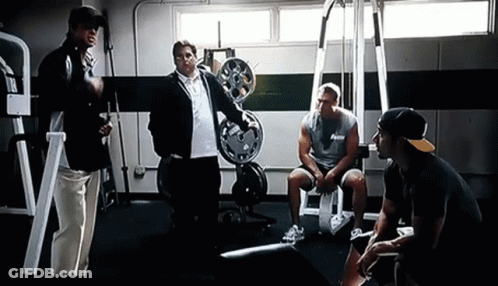 A Group Of Men Are Standing And Sitting In A Gym Talking To Each Other GIF