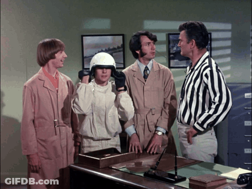 A Group Of Men Are Standing Around A Desk And One Of Them Is Wearing A White Helmet GIF