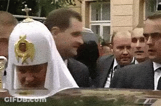 A Group Of Men Are Standing Around A Man Wearing A White Hat With A Cross On Top . GIF