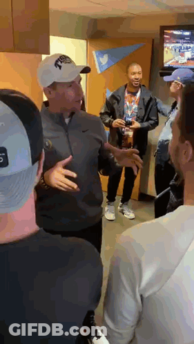A Group Of Men Are Standing In A Room Talking To Each Other . One Of The Men Is Wearing A Nike Hat . GIF