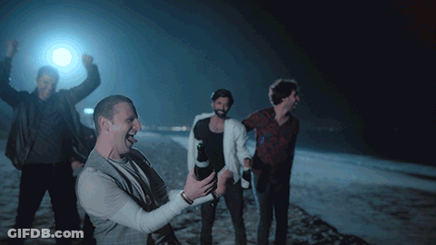 A Group Of Men Are Standing On A Beach At Night And One Of Them Is Holding A Bottle Of Beer GIF