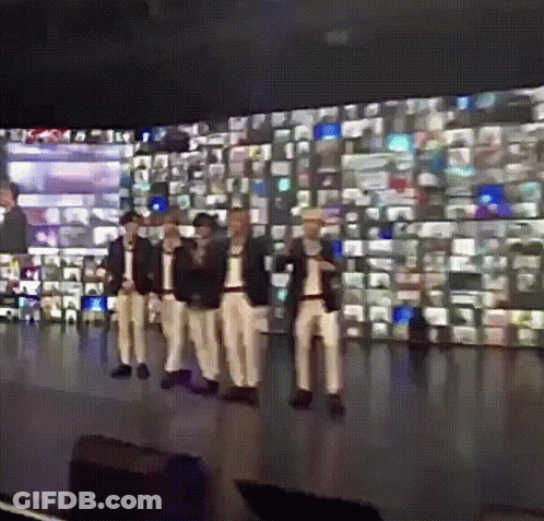 A Group Of Men Are Standing On A Stage In Front Of A Wall Of Television Screens GIF