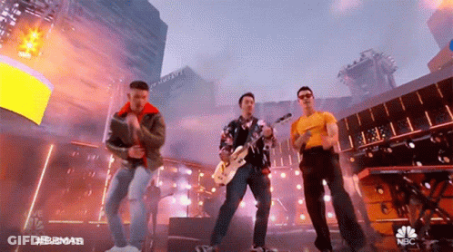 A Group Of Men Are Standing On A Stage Playing Instruments And Singing Into Microphones . GIF